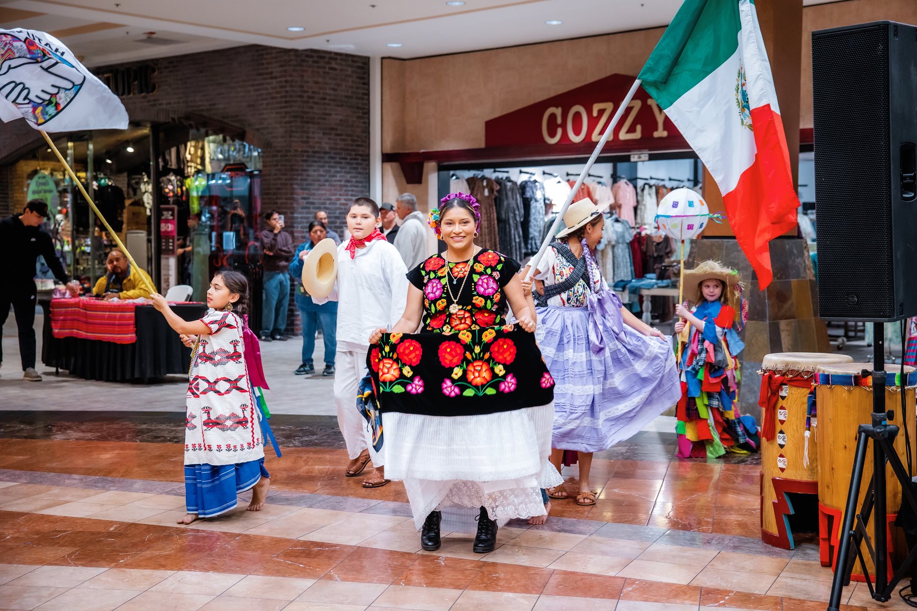 Participantes con vestimenta colorida están frente al escenario con una gran bandera de México