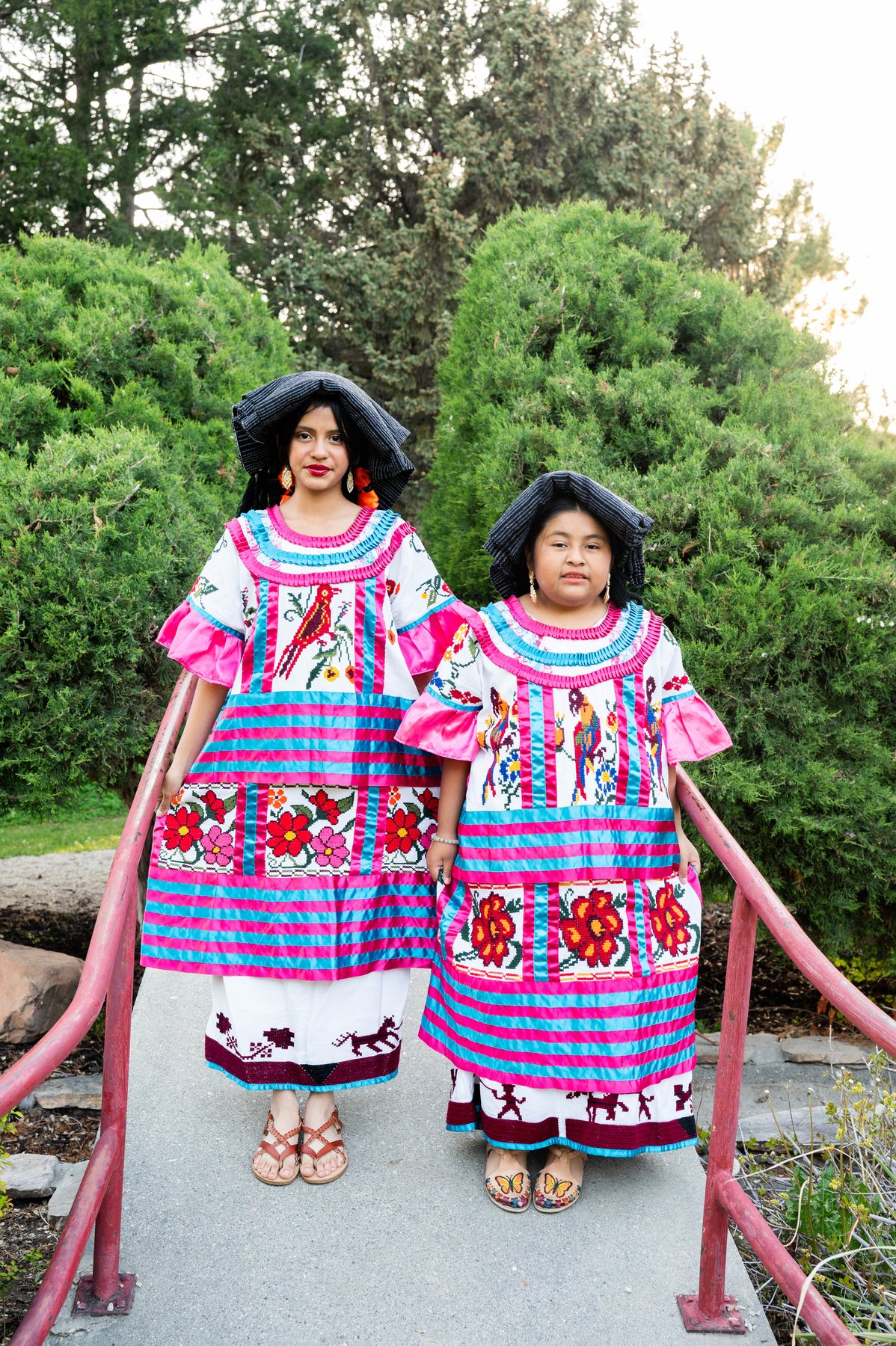 Dos participantes de Oaxaca en Utah posan con vestimenta bordada de colores sobre un puente de jardín