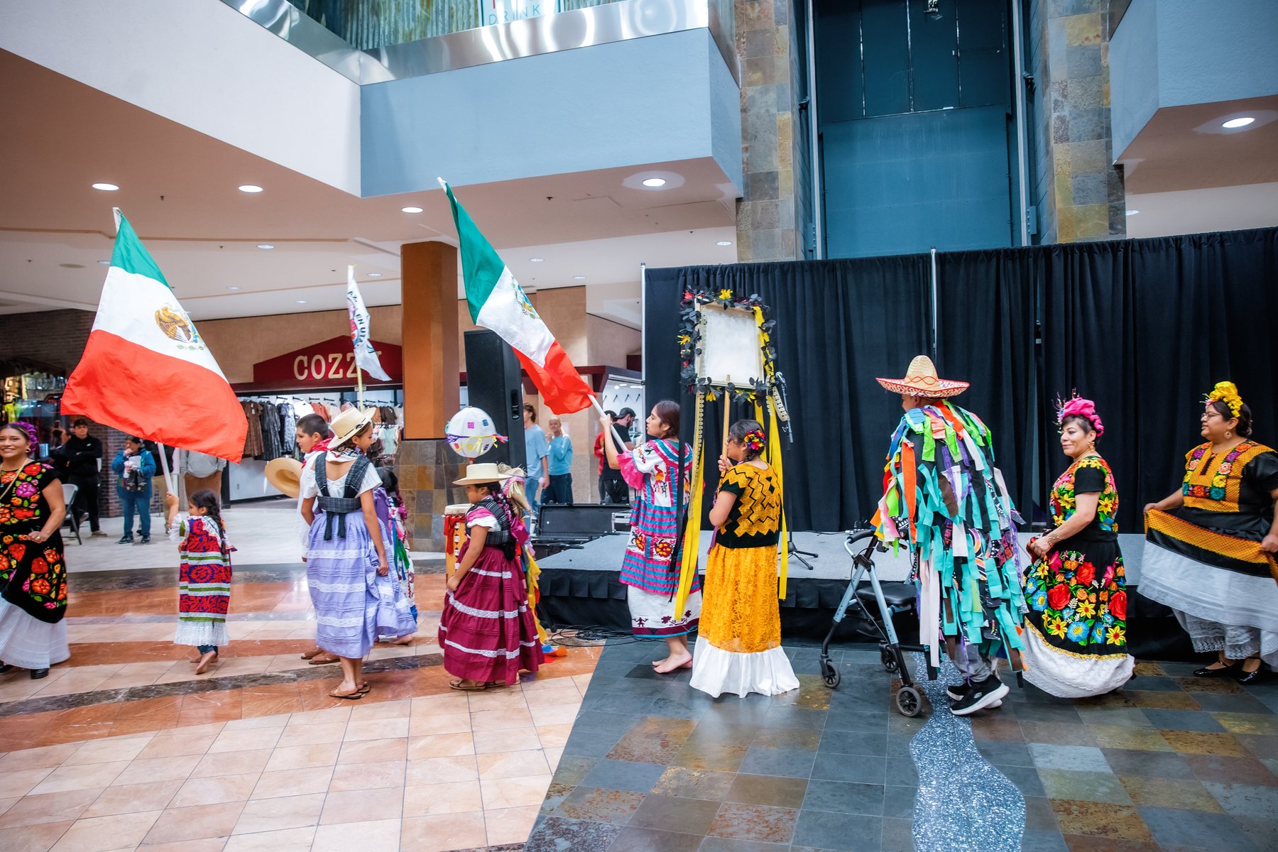 Una fila de participantes con vestimenta colorida está con banderas de México junto al escenario