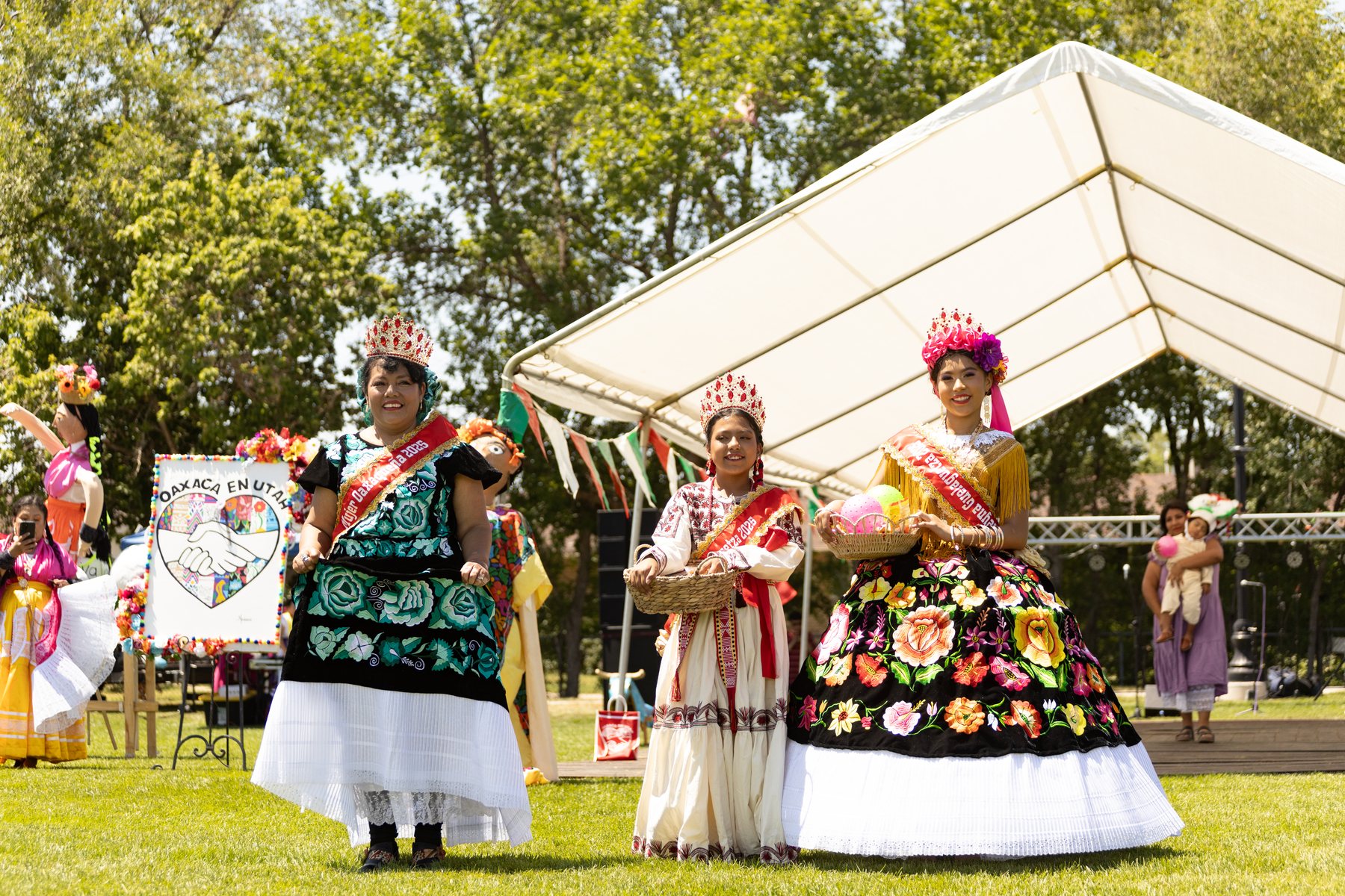 Tres participantes con vestidos bordados están juntas junto al escenario