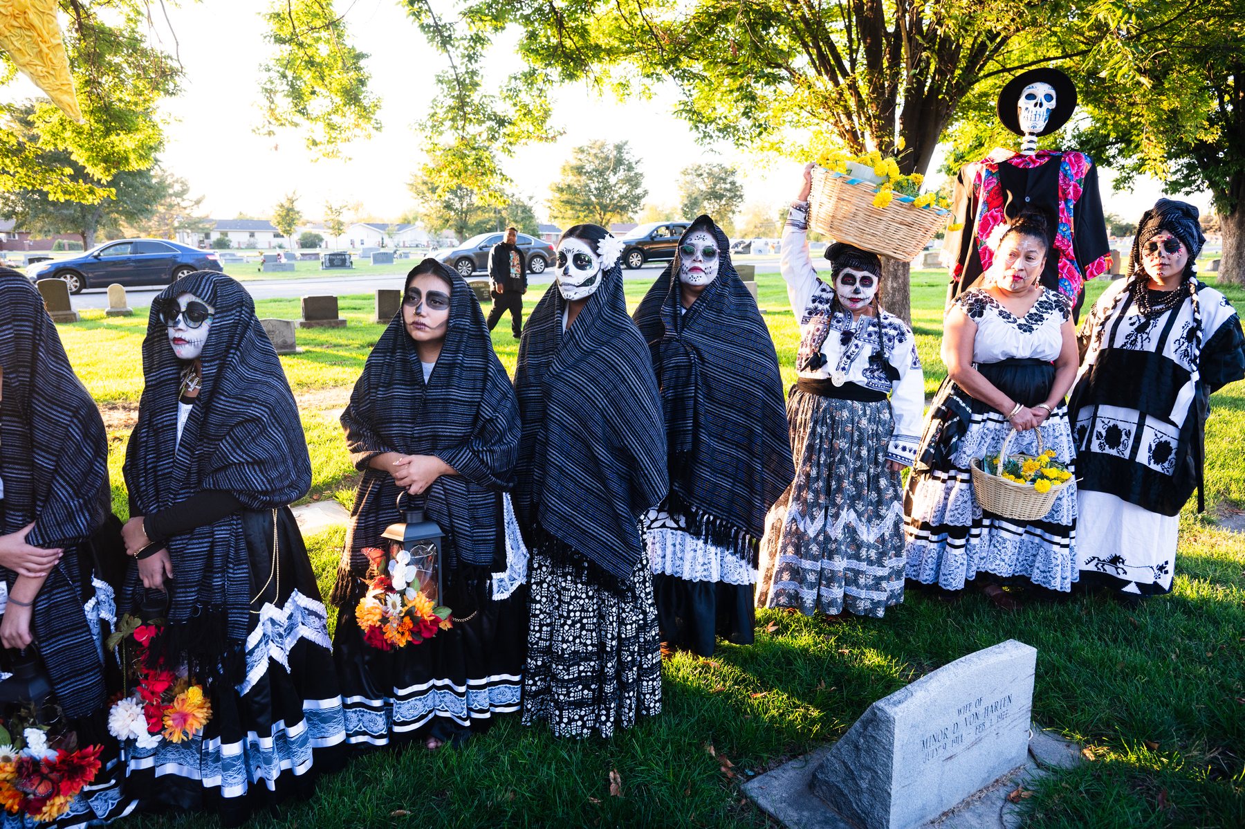 Un grupo con vestimenta negra y blanca se reúne junto a una tumba y una figura alta de esqueleto