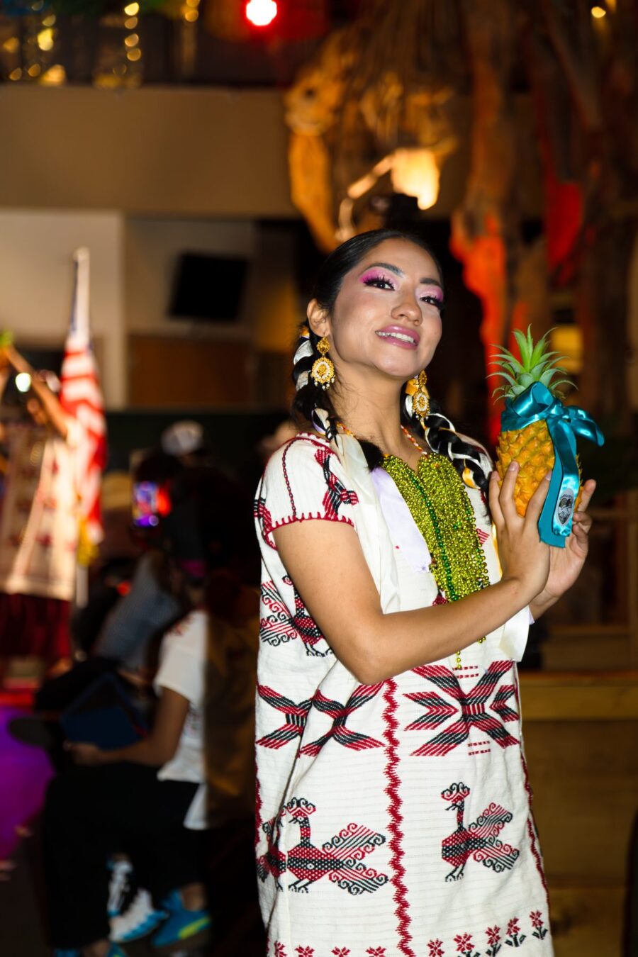 A model in a white woven garment with red and black designs holds a ribbon-decorated pineapple