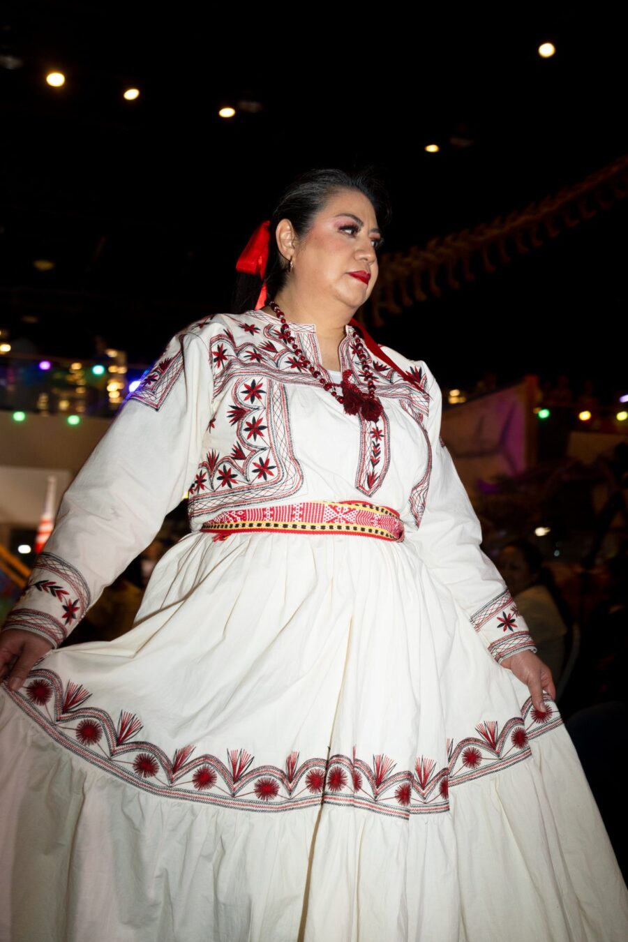 A model displays a white dress with red and black embroidery beneath the runway lights